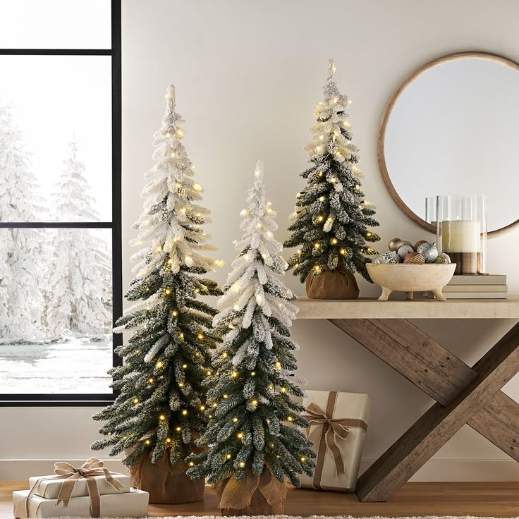 Artificial snow-covered Christmas trees with lights on a wooden table, surrounded by festive decor in a cozy, wintry room setting.