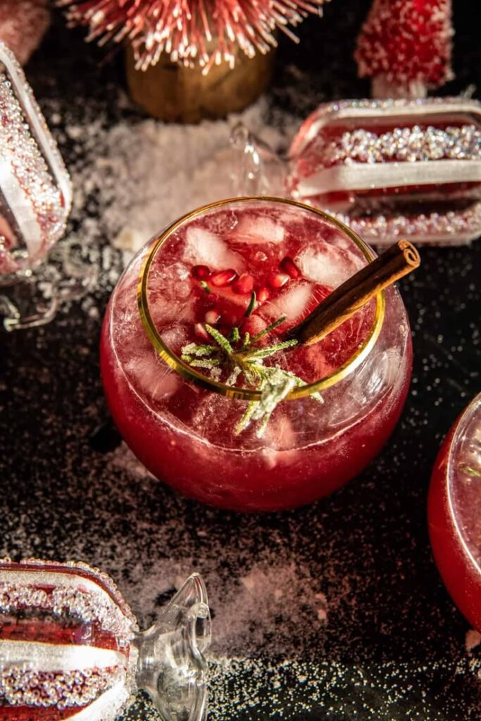 Festive pomegranate cocktail with cinnamon stick and ice, surrounded by holiday decorations and glass ornaments.