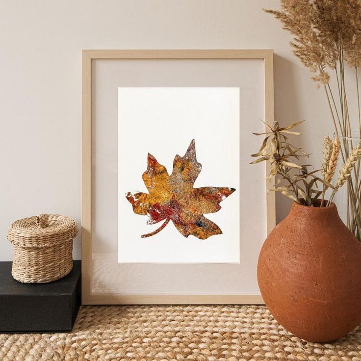 Framed autumn leaf art on a textured surface, surrounded by rustic decor including a woven basket and dried floral arrangement.