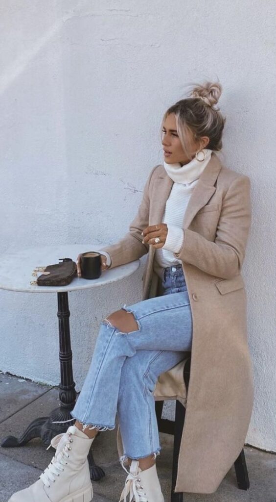 Woman in stylish beige coat and ripped jeans, holding a coffee cup, sitting at a table outdoors. Fashionable winter look.