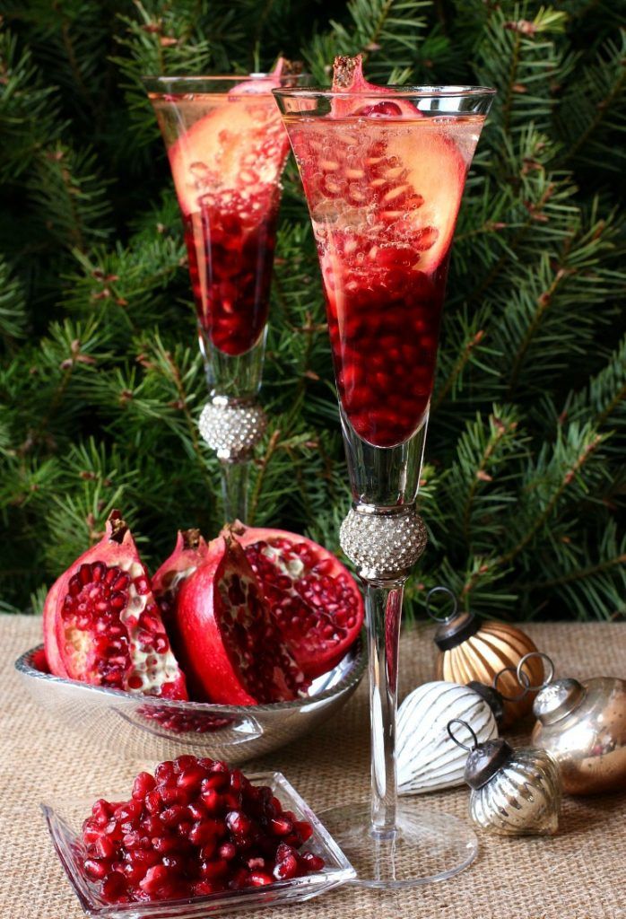 Festive pomegranate cocktail in elegant glasses with sliced fruit, surrounded by holiday ornaments on a burlap table.