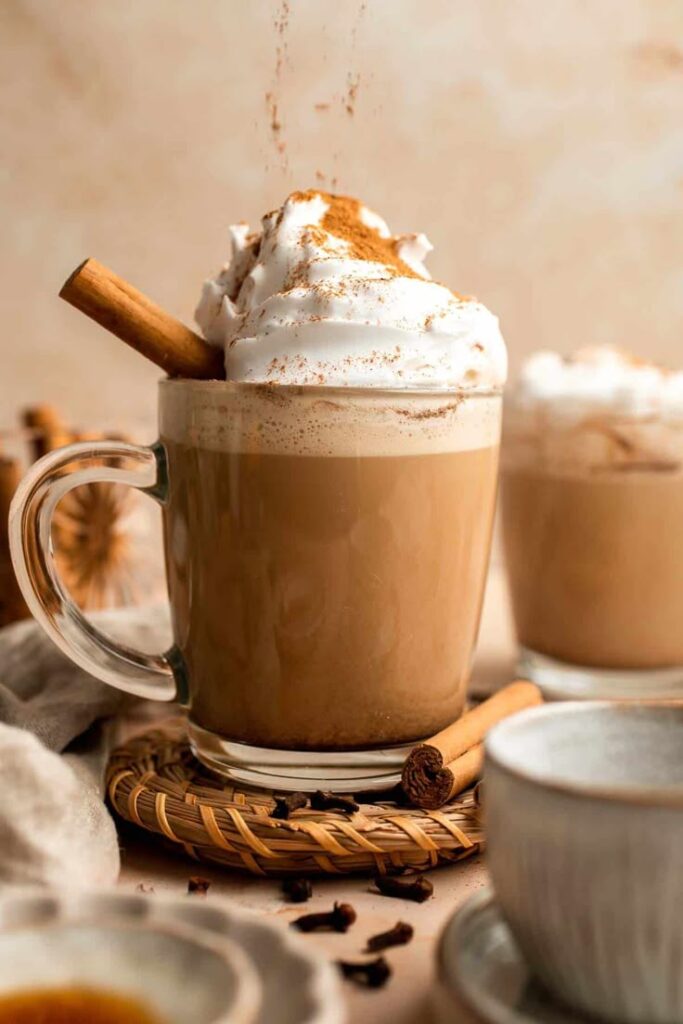 Creamy latte topped with whipped cream, cinnamon stick, and sprinkled spices, served in a clear mug for a cozy autumn treat.