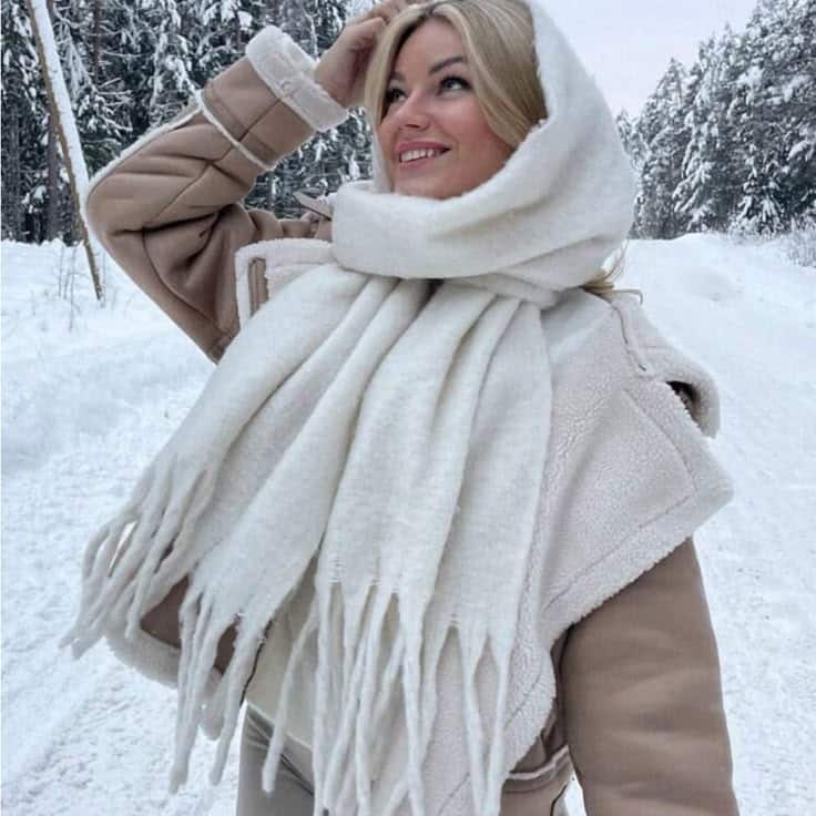 Woman in winter attire smiles in snowy forest, wearing beige coat and white scarf. Winter fashion photography.