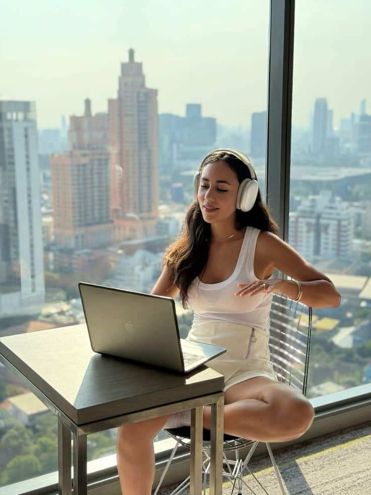 Woman enjoying music with headphones, working on a laptop in a bright high-rise room with a city view.