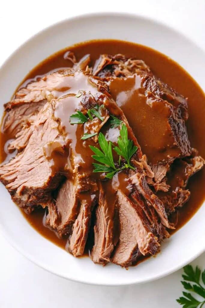 Tender roast beef covered in rich gravy, garnished with fresh parsley on a white plate. Perfect for a hearty meal.