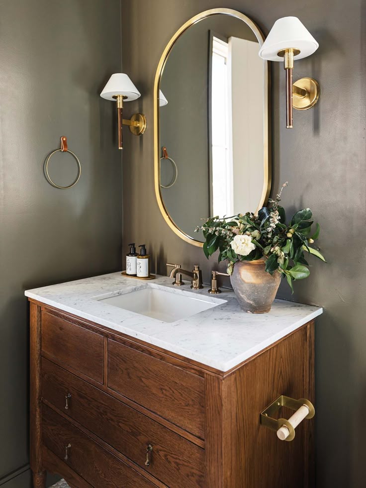 Elegant bathroom with a wooden vanity, marble countertop, oval mirror, and stylish wall sconces. Greenery accents the serene space.