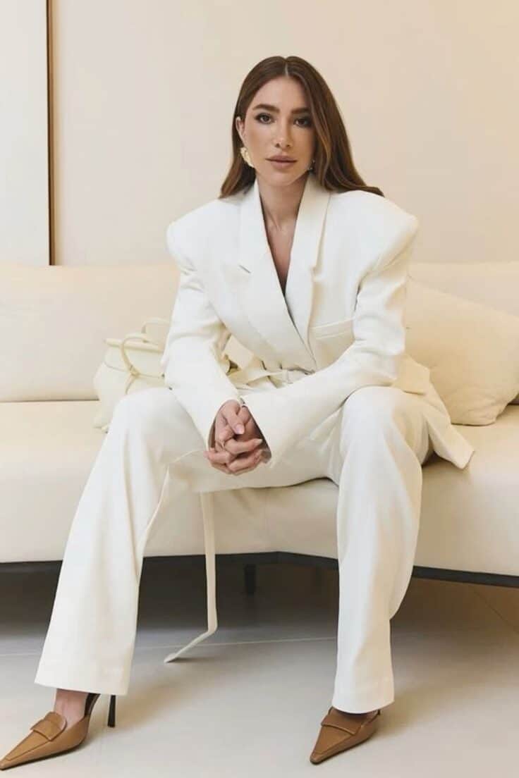 Woman in white suit sits confidently on a sofa, wearing brown shoes and gold earrings in a minimalist room. Fashionable elegance.