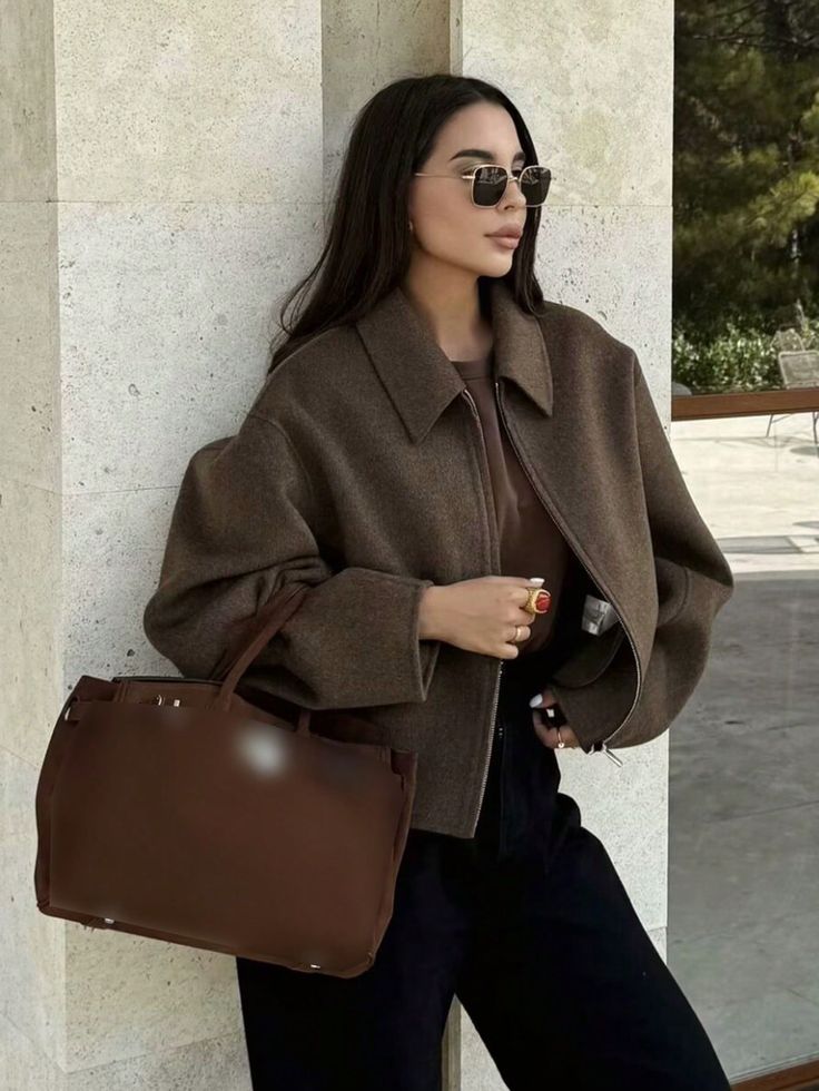 Fashionable woman in sunglasses, brown coat, and large handbag standing by a stone wall in outdoor setting. Chic and stylish look.