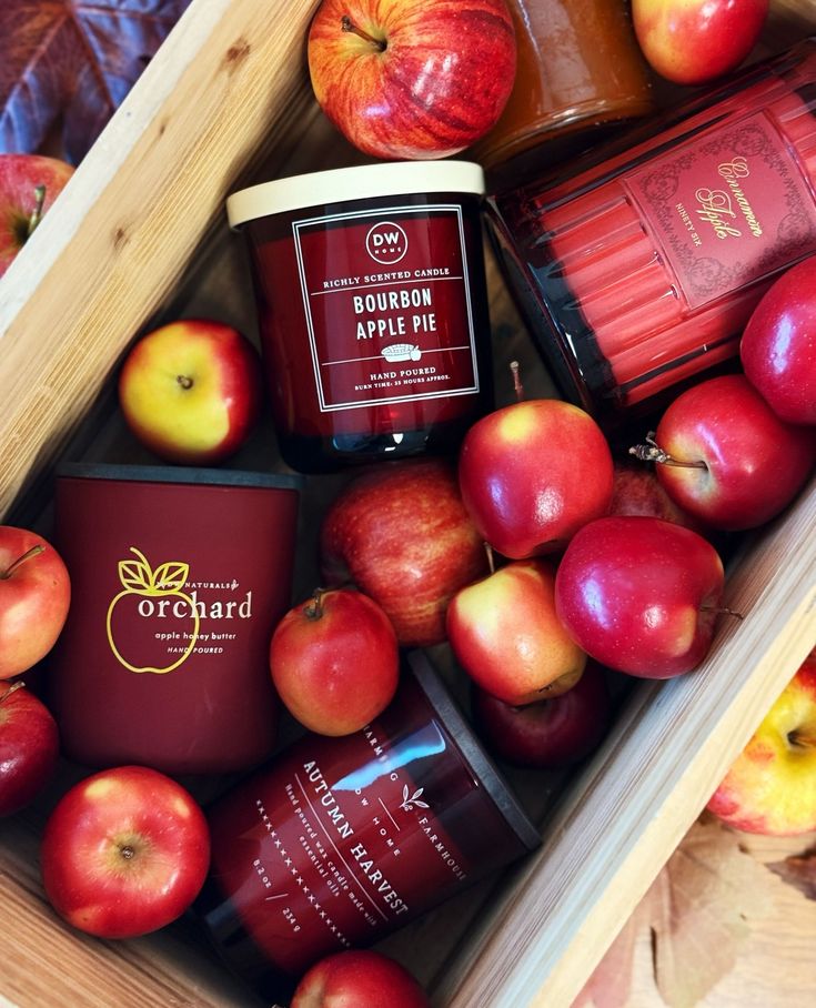 Autumn-themed candles with red apples in a wooden crate, featuring scents like Bourbon Apple Pie and Cinnamon Apple. Cozy fall decor.