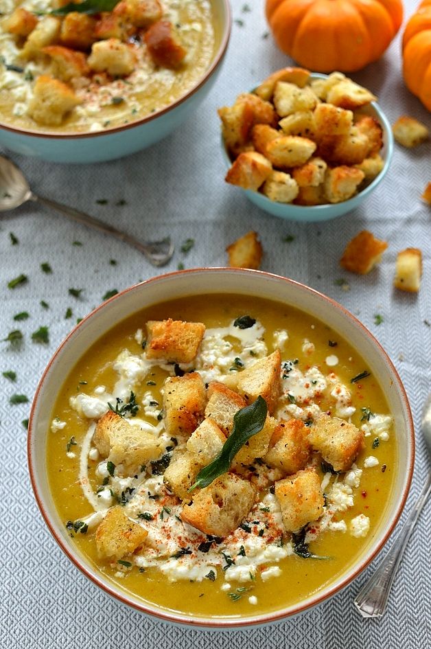 Bowl of creamy pumpkin soup topped with croutons, herbs, and cream, next to mini pumpkins and extra croutons on a gray cloth.