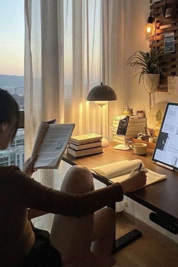Cozy study nook with books, lamp, and tablet; person reading with warm evening light. Relaxed, productive atmosphere.