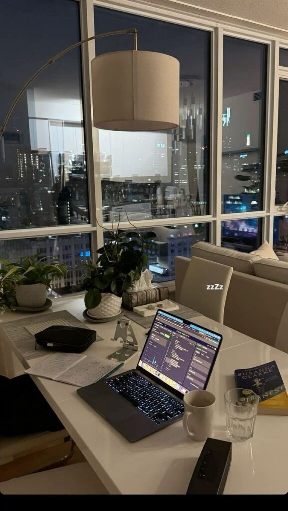 Cozy nighttime workspace with a laptop, coding screen, and city skyline view through large windows.
