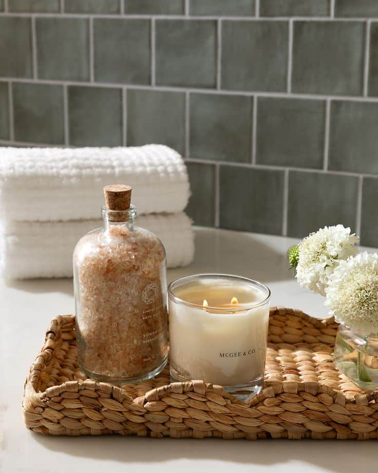 Cozy spa setup with a lit candle, bath salts, white towels, and flowers on a woven tray in a serene bathroom setting.