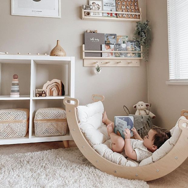 Cozy baby room with a child reading in a wooden rocker, surrounded by books, toys, and a plush rug. Calm, minimalist decor.