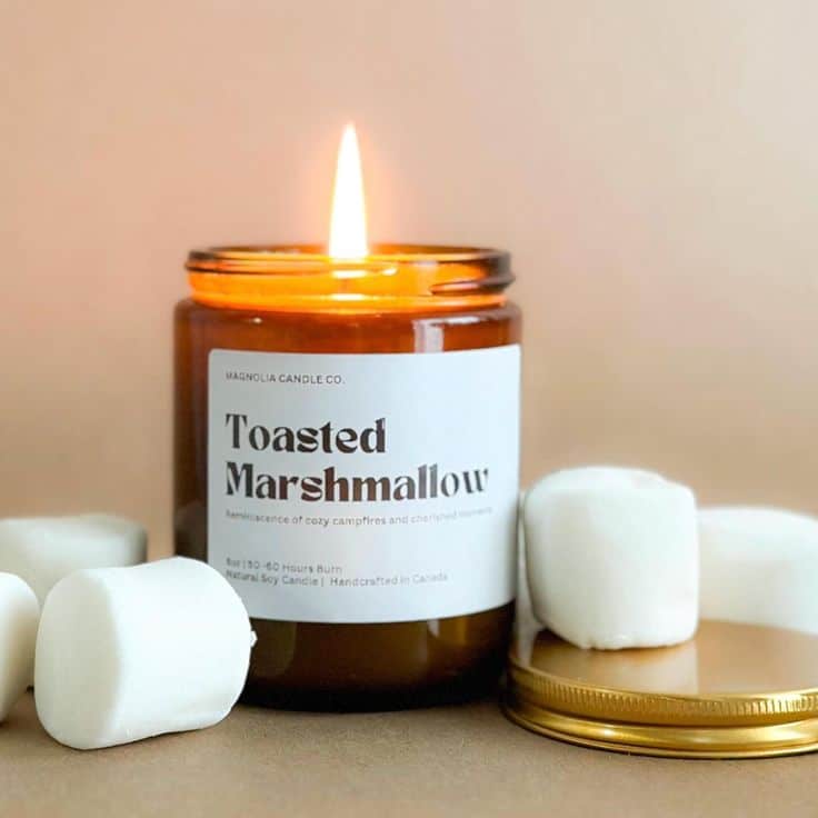 Toasted marshmallow-scented candle burning in amber jar, surrounded by marshmallows, creating a cozy campfire vibe.
