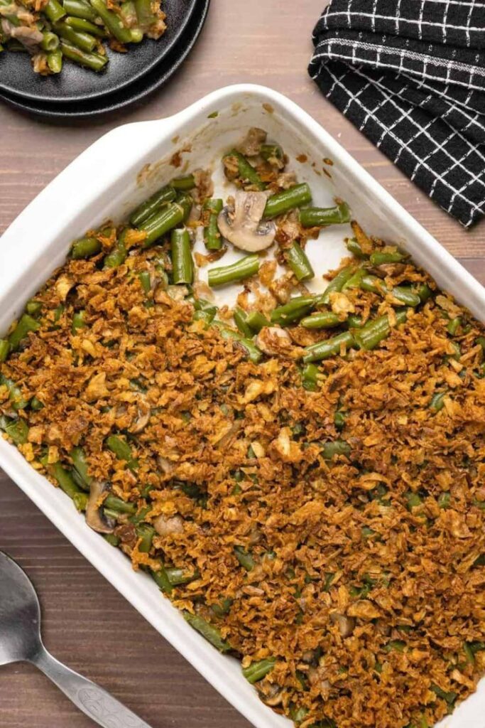Green bean casserole topped with crispy onions in a white dish, accompanied by a serving on a black plate. Perfect holiday side dish.
