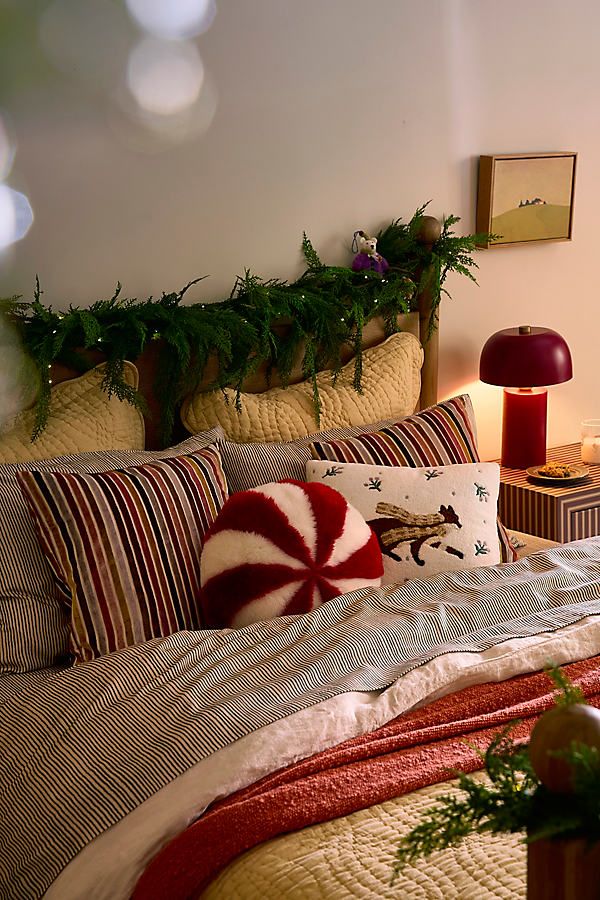 Cozy bedroom with festive decor, featuring striped bedding, plush pillows, greenery on headboard, and a warm red lamp.
