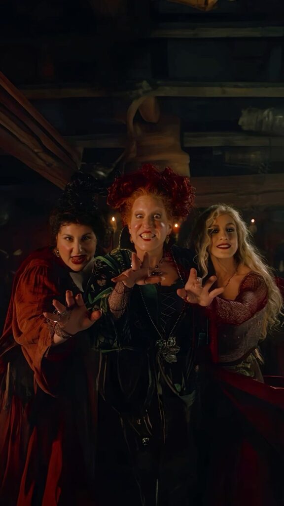 Three witches casting a spell, dressed in vibrant costumes, reaching out with dramatic expressions in a dimly lit room.
