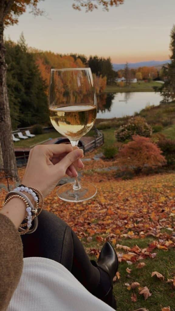 Glass of white wine held over a scenic autumn landscape with colorful leaves, a serene pond, and distant trees at sunset.