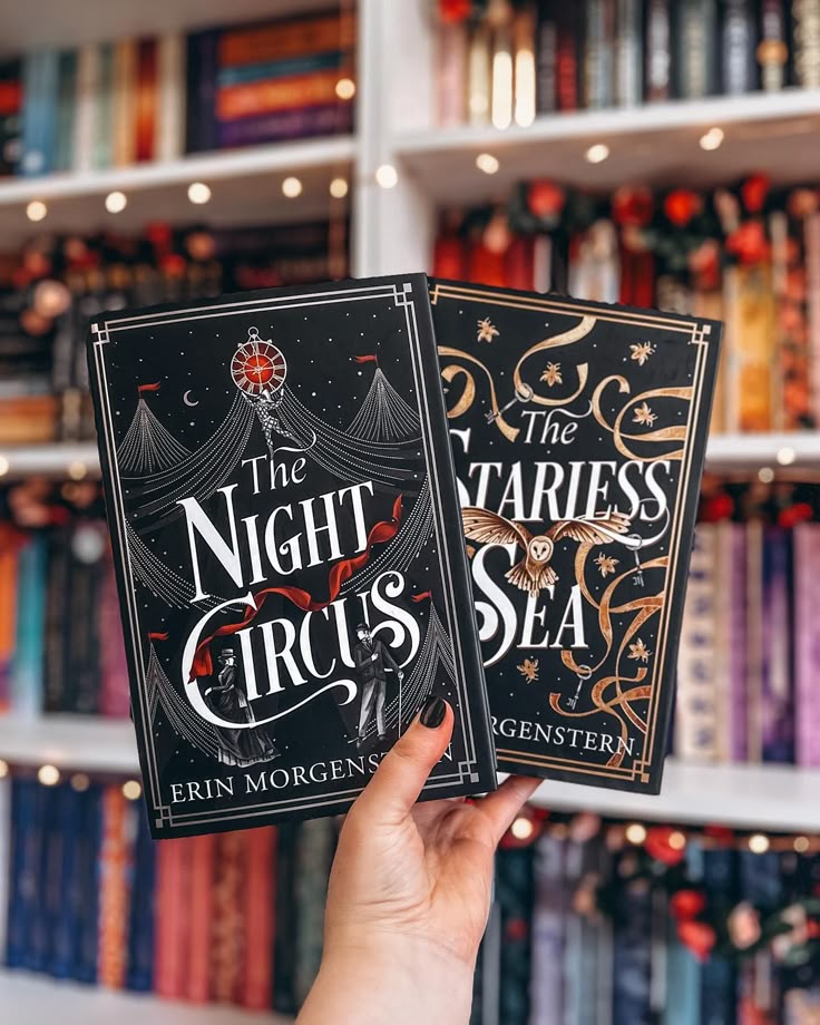 A hand holds two books, The Night Circus and The Starless Sea, in front of a colorful bookshelf.