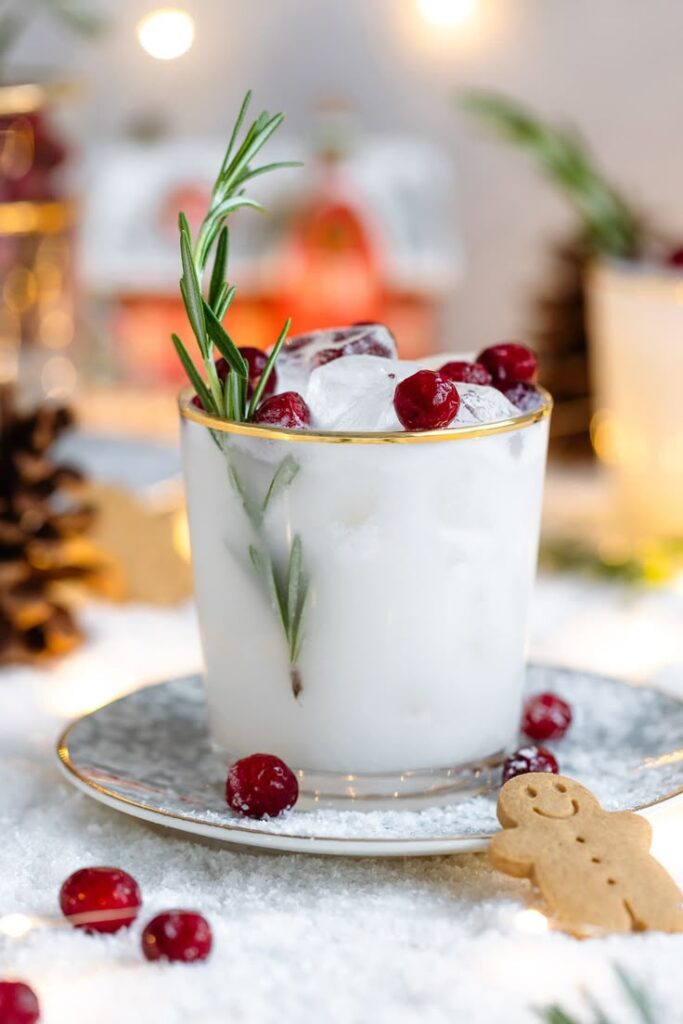 Festive holiday cocktail with rosemary, cranberries, and ice in a glass, surrounded by snow and gingerbread cookie.