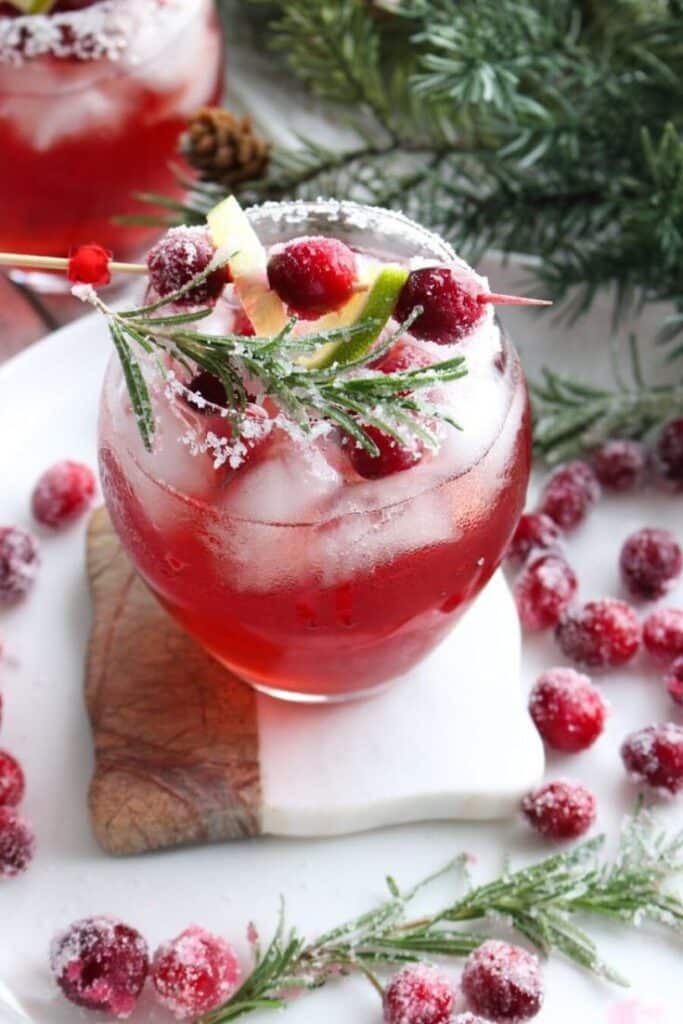 Festive cranberry cocktail with rosemary garnish, surrounded by sugared cranberries, perfect for holiday celebrations.