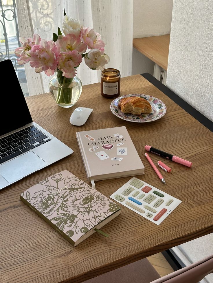 Cozy workspace with laptop, pink flowers, notebooks, pens, and croissant. Ideal for productivity and inspiration.