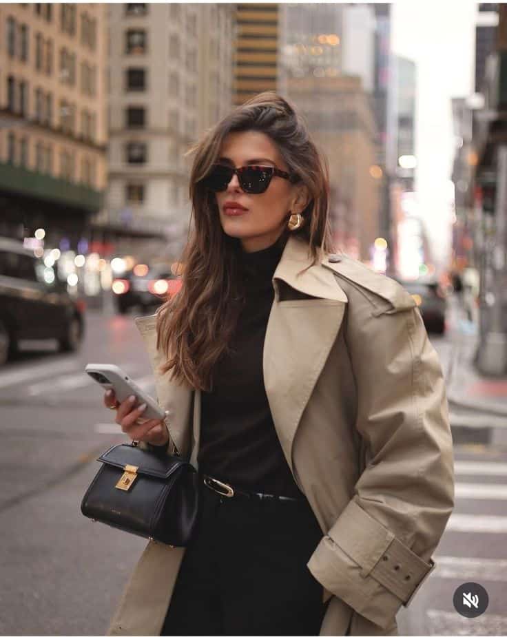 Woman in trench coat with phone and handbag, standing in urban street setting, wearing sunglasses and stylish earrings.