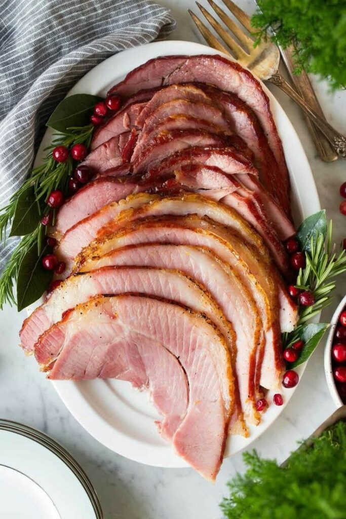 Sliced holiday ham garnished with rosemary and cranberries, served on a white platter with festive decor.