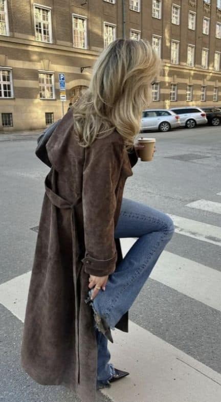Woman in a brown coat and jeans holding coffee, stepping on a crosswalk in an urban setting.
