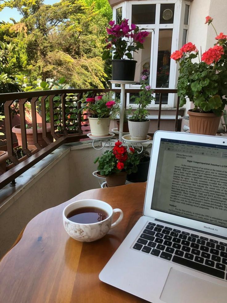 Laptop and coffee on a wooden table in a balcony garden, surrounded by colorful flowers, with a peaceful outdoor view.