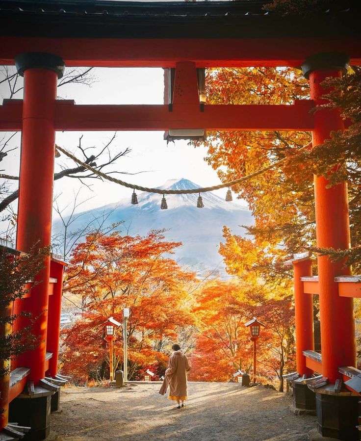 Red torii gate framing Mount Fuji, surrounded by vibrant autumn leaves in Japan. Person walking below. Scenic and serene view.