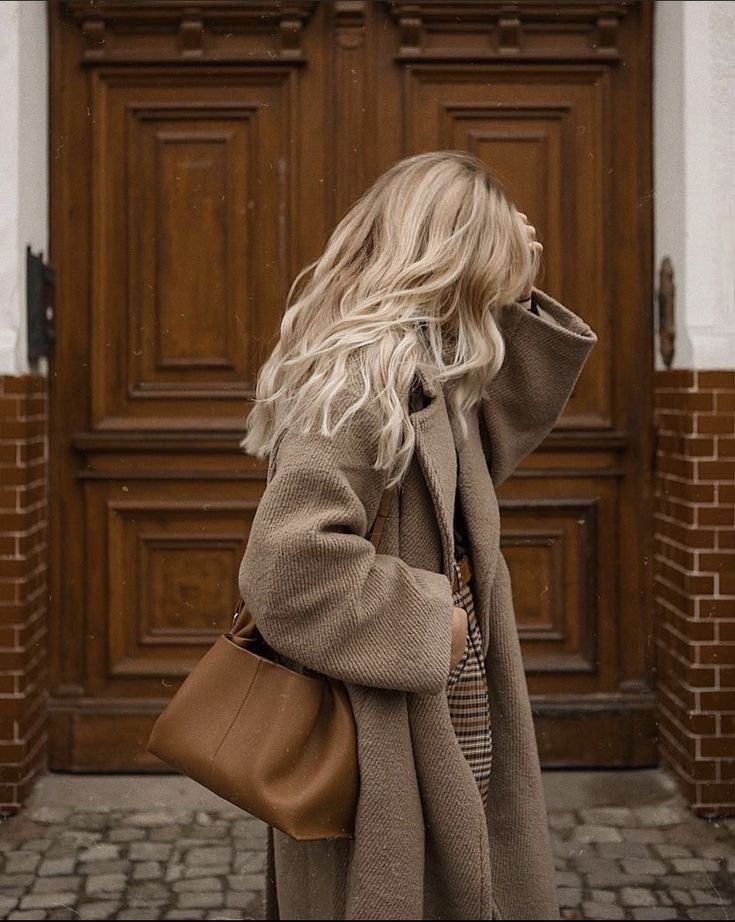 Woman with long blonde hair in a cozy brown coat poses stylishly by wooden doors, carrying a brown leather bag. Fashionable autumn look.