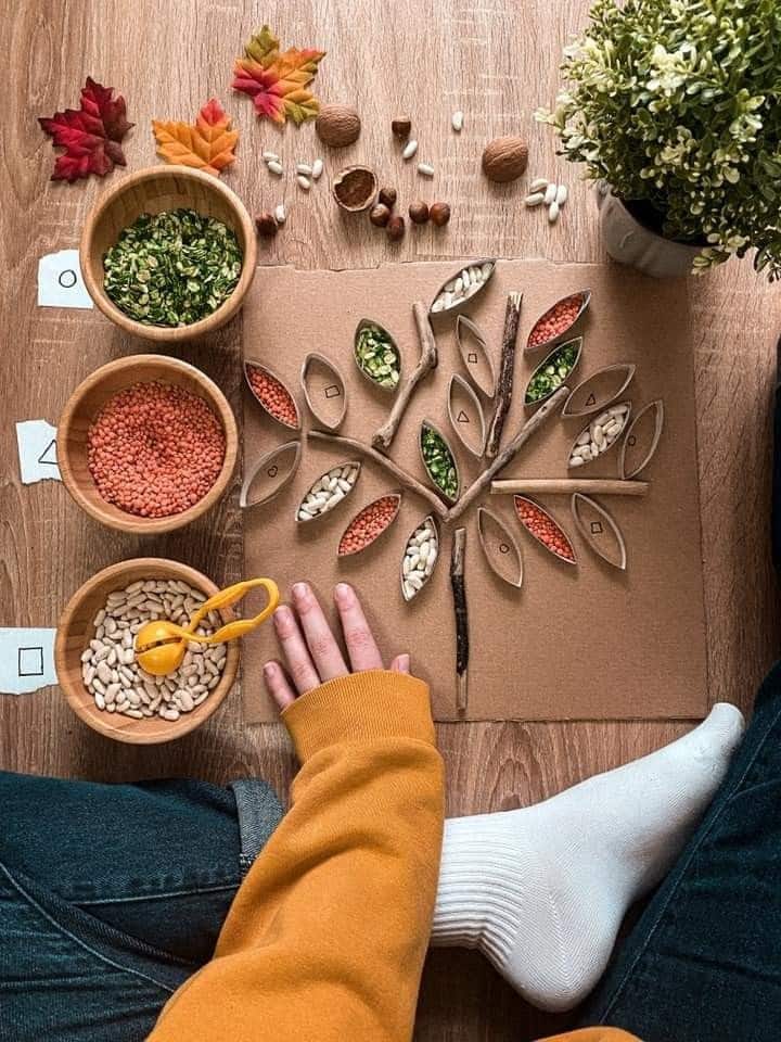 Creative autumn craft: cardboard tree design with colorful beans and seeds, surrounded by bowls of ingredients on a wooden floor.