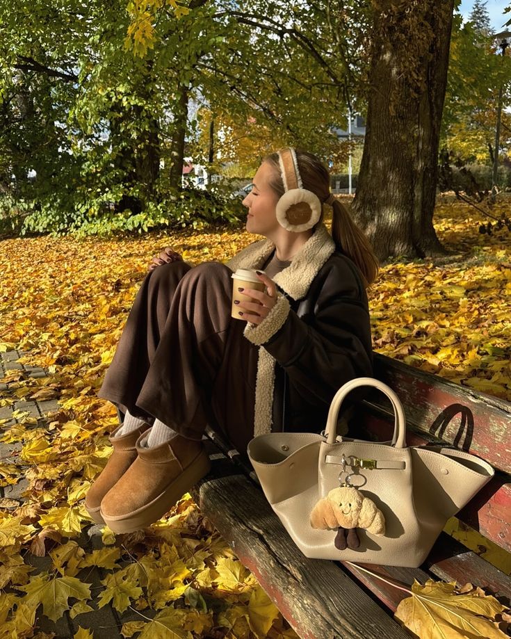Woman in cozy fall attire on a bench, holding coffee, surrounded by autumn leaves in a park setting.