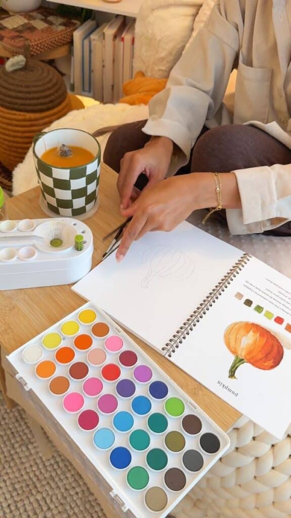 Person sketching a pumpkin with watercolors and a checked mug nearby on a cozy table setting.