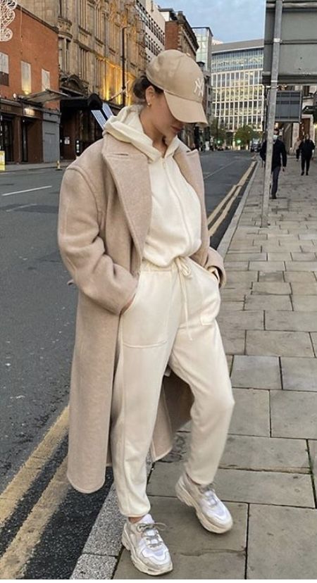 Stylish woman in beige coat, white hoodie, joggers, and sneakers, walking urban street. Neutral cap complements chic, casual look.