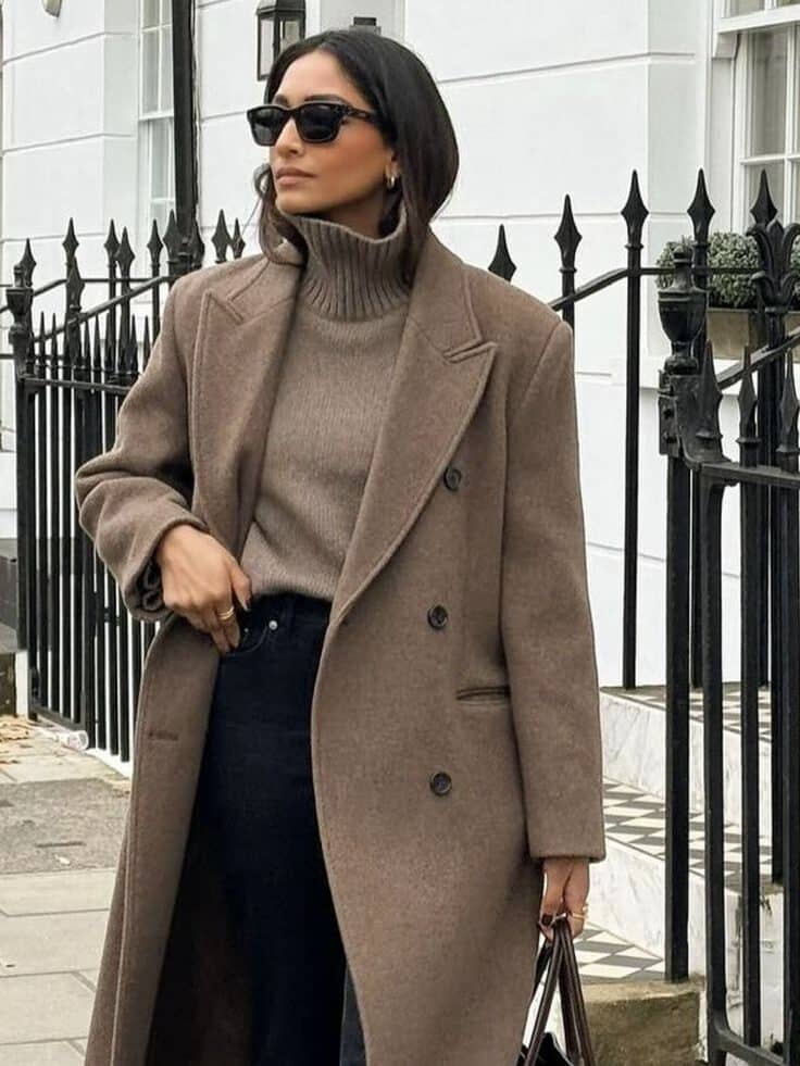 Stylish woman in a long brown coat and sunglasses, walking on a city street with a large handbag, showcasing fall fashion.