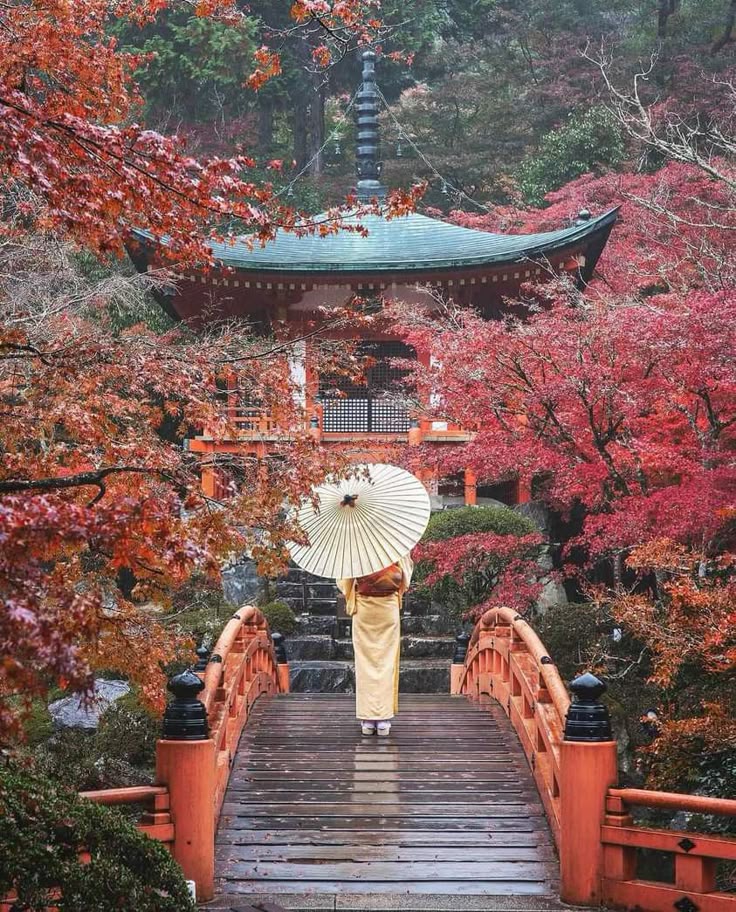 A person in traditional attire with an umbrella on a red bridge amid vibrant autumn foliage, leading to a pagoda in Japan.