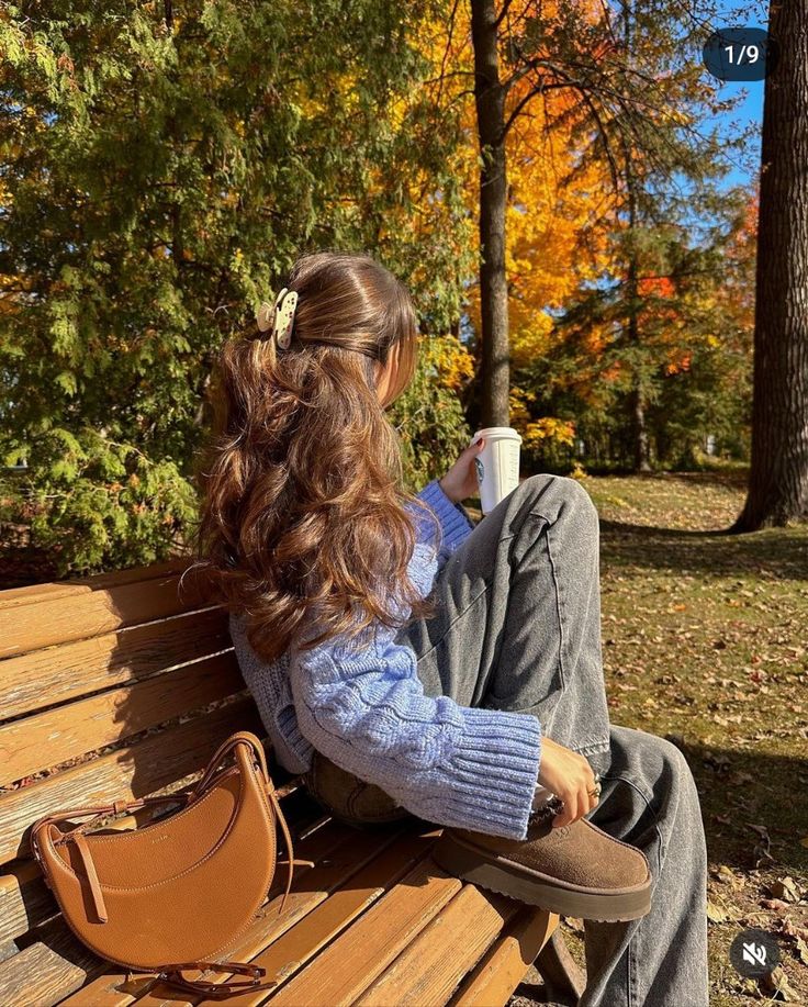 Woman in a blue sweater sipping a drink on a park bench in autumn, surrounded by colorful trees and sunlight.