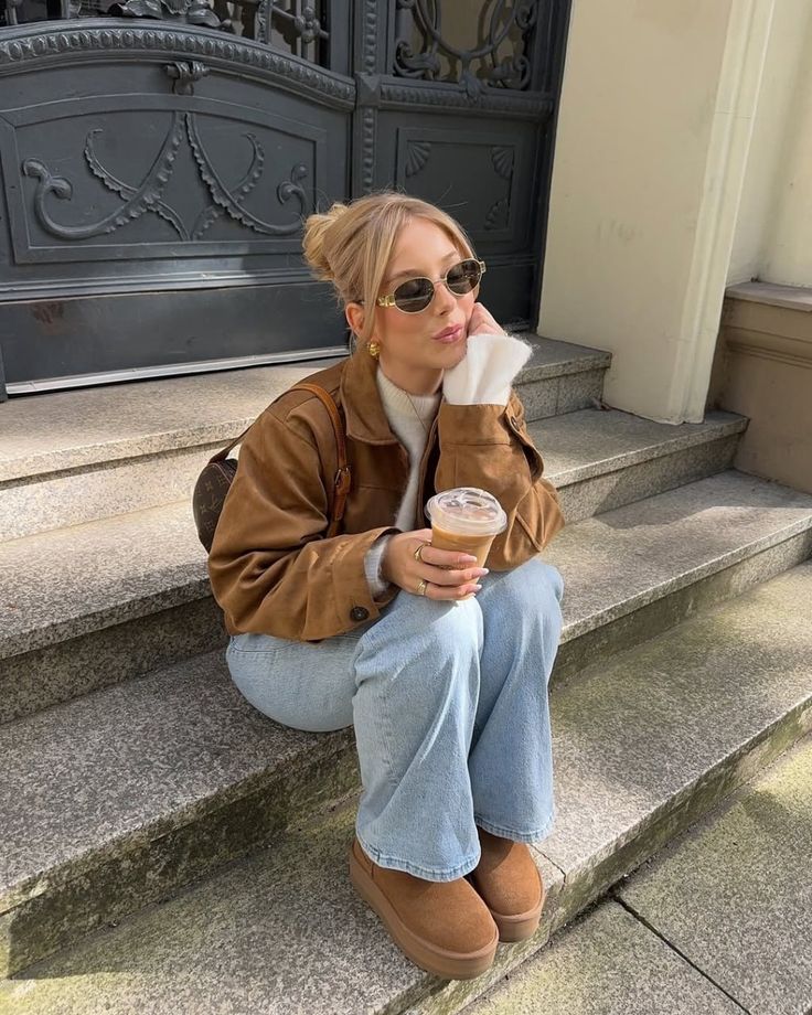 Woman sitting on steps, wearing sunglasses and a brown jacket, holding iced coffee. Casual autumn fashion.