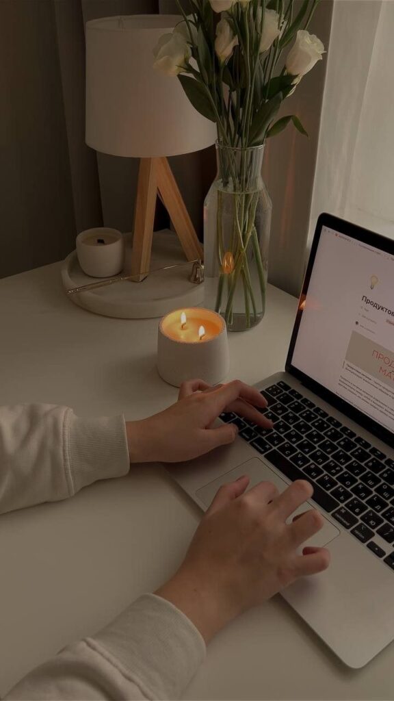 Person typing on a laptop in a cozy workspace with flowers, candle, and lamp for a calming ambiance.