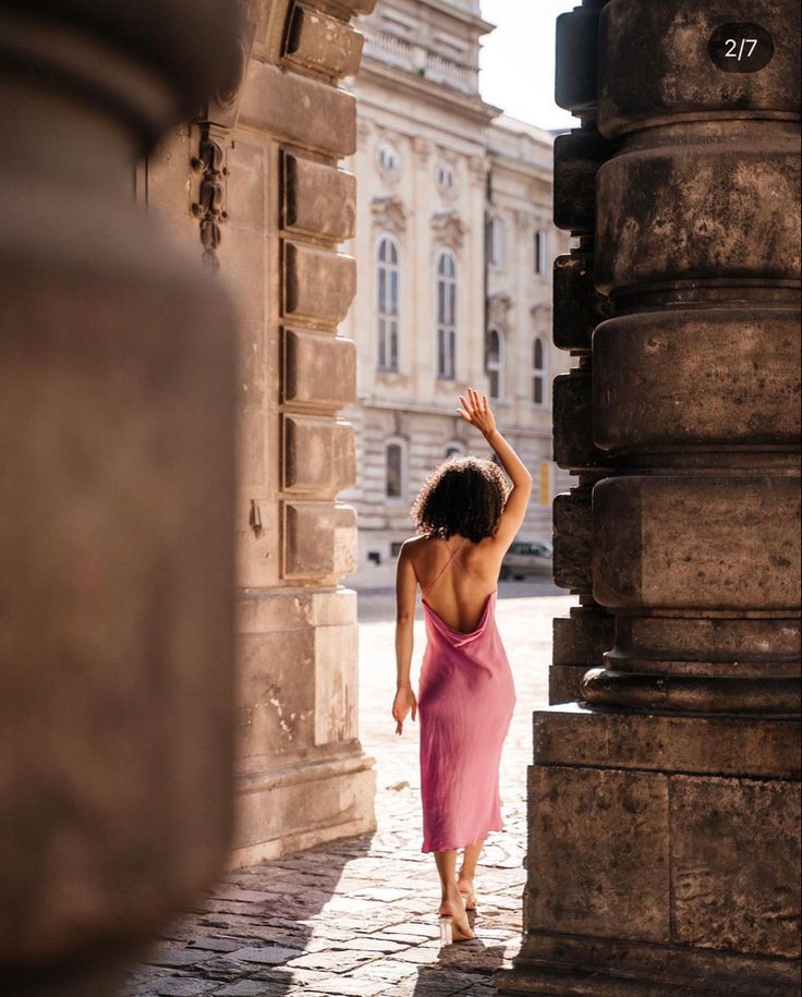 Woman in pink dress walking through historic stone archway, hand raised, with sunlight casting shadows. Urban elegance and architecture.