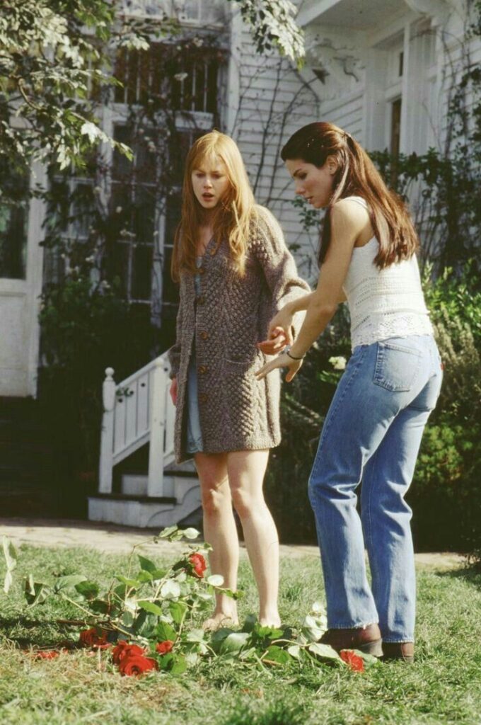 Two women stand in a garden near a white house, examining roses on the ground, wearing casual clothing.