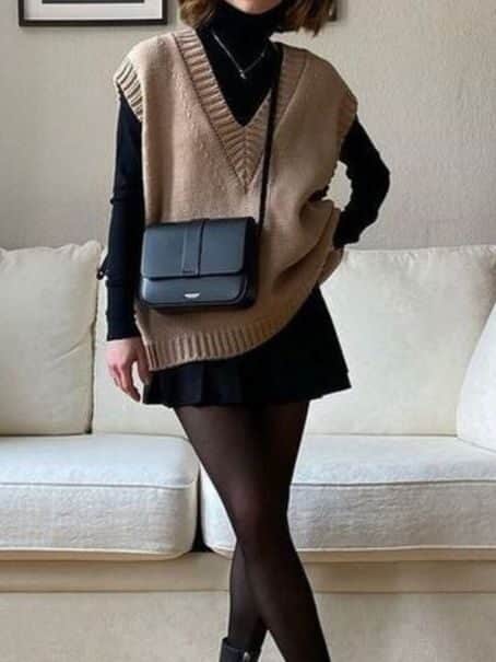 Fashionable woman in a beige vest and black boots, posing stylishly in a modern living room setting.
