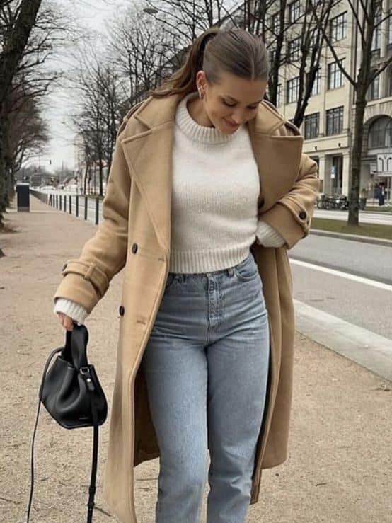 Woman in a stylish beige coat, white sweater, jeans, and sneakers holding a black bag, walking on a city sidewalk.
