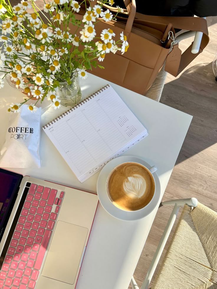 Bright workspace with a latte, planner, laptop, flowers, and a bag on a sunny day. Perfect setup for productivity and relaxation.