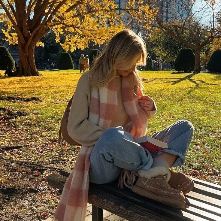 Woman reading a book on a sunny autumn day, surrounded by golden leaves, wearing cozy clothes and sitting on a park bench.