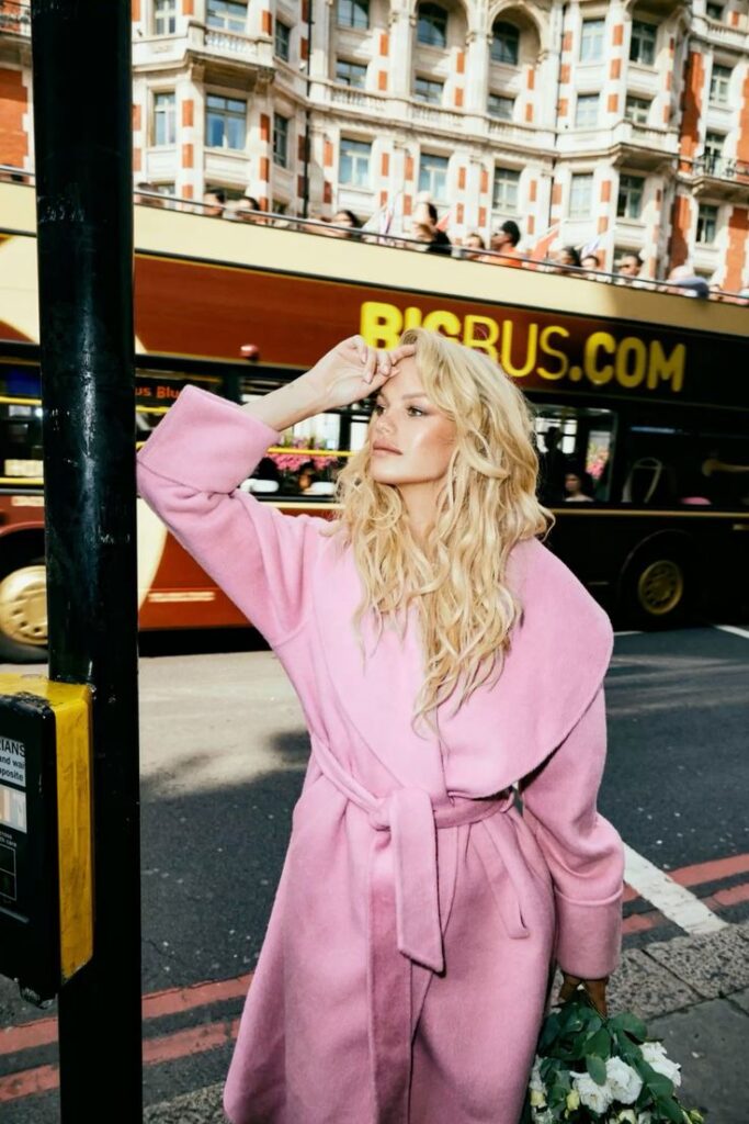 Woman in a pink coat stands on a city street, holding flowers, with a sightseeing bus and historic building in the background.