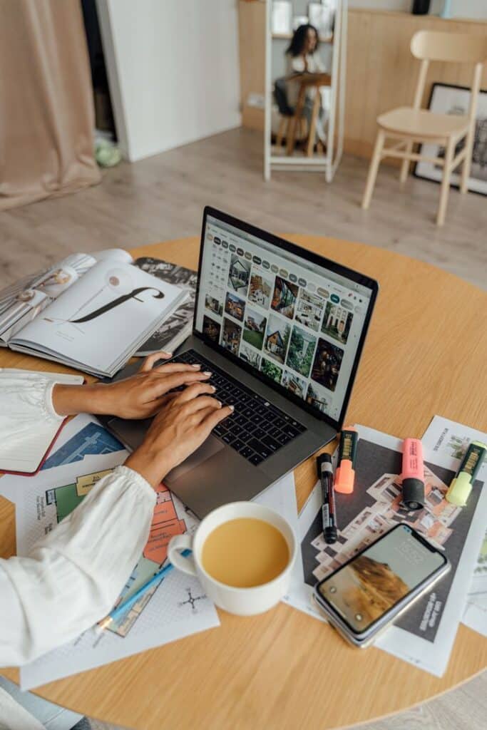 Person browsing design ideas on a laptop, surrounded by papers, magazines, and a cup of coffee on a wooden table.